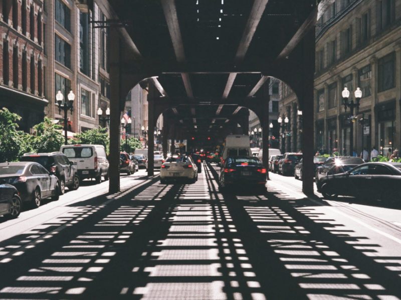 cars drive through the Chicago loop, in the shadows of overhead transit rail lines