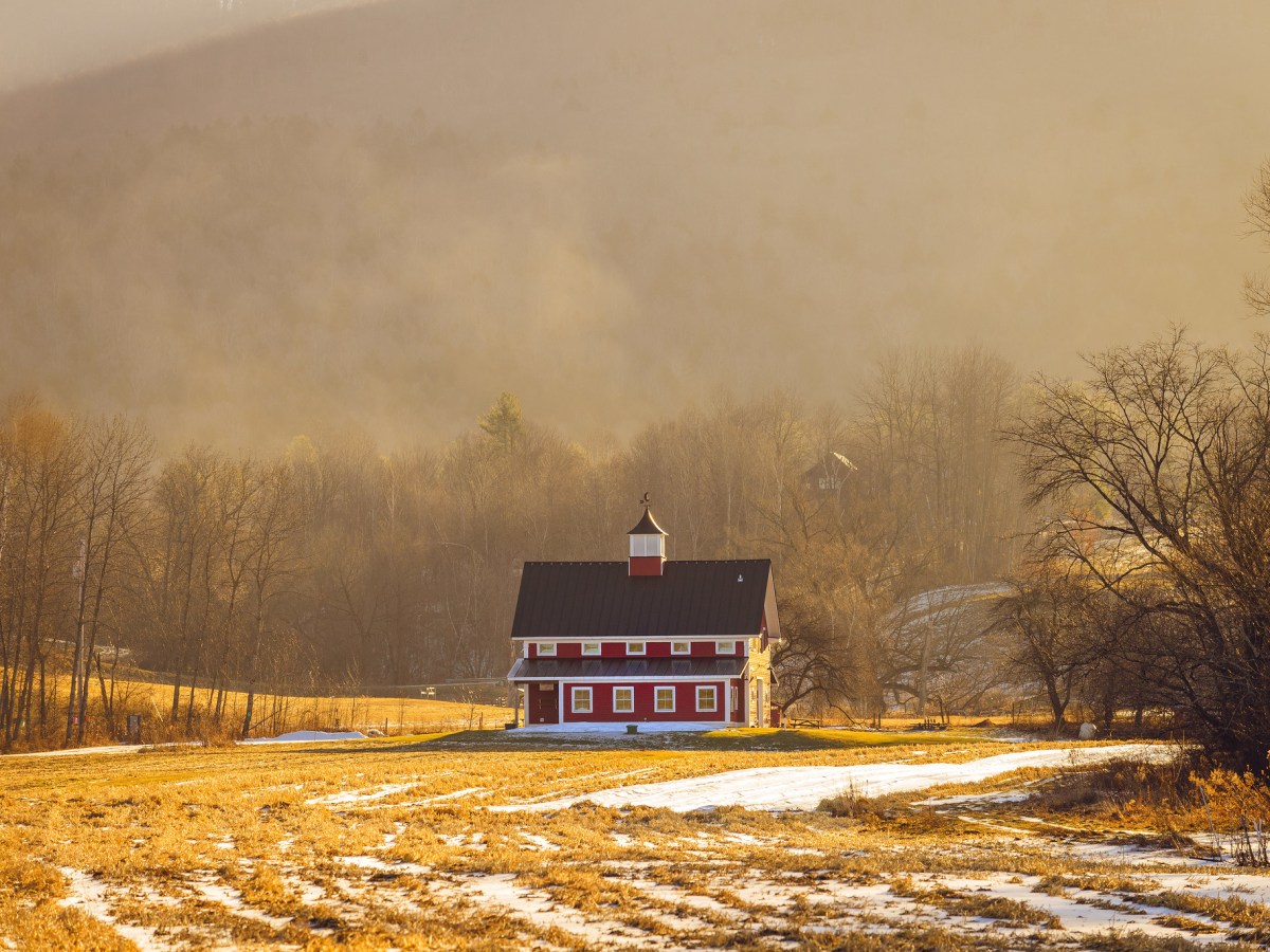 A large red barn sits in a golden field streaked with just a bit of snow