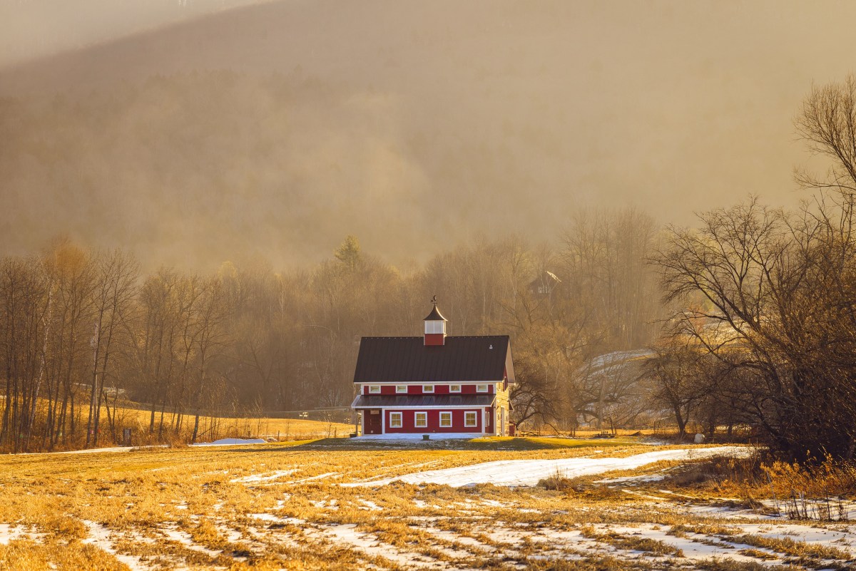 A large red barn sits in a golden field streaked with just a bit of snow