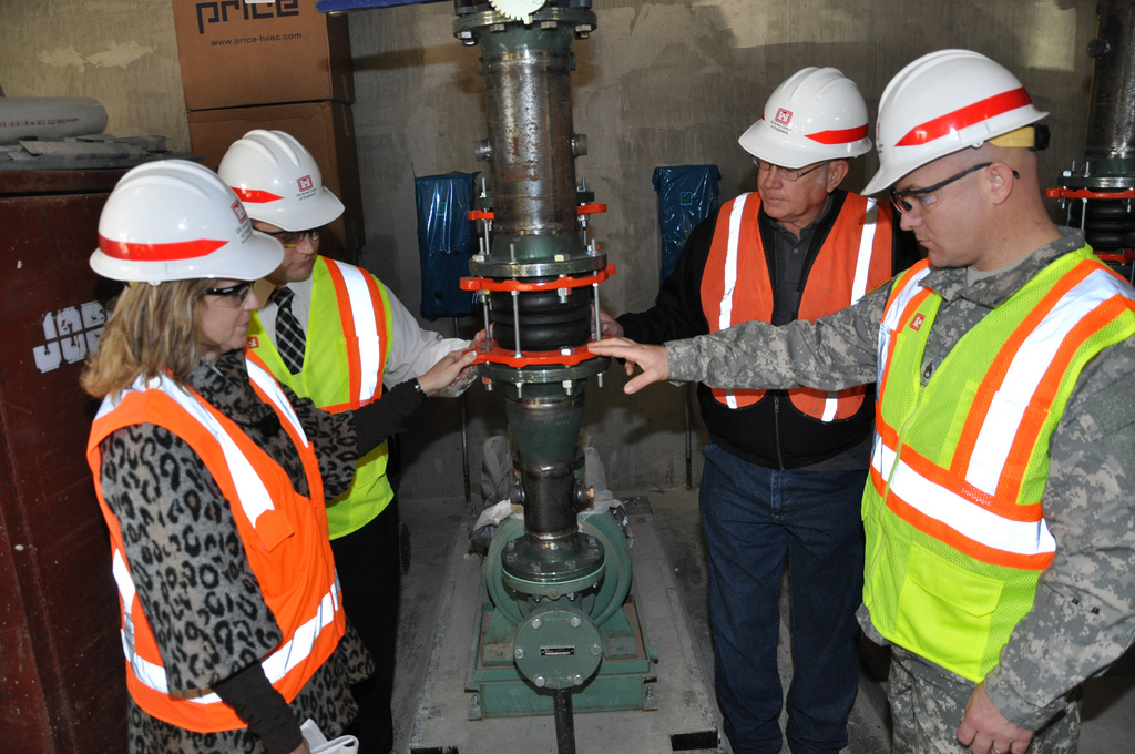 Four people in hard hats and neon reflective vests observe a metal device that is a ground-source heat pump.