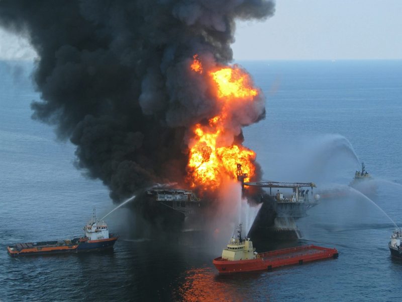 Coast guard boats gather around a massive fire in the ocean spewing black smoke, trying to put out the flames.