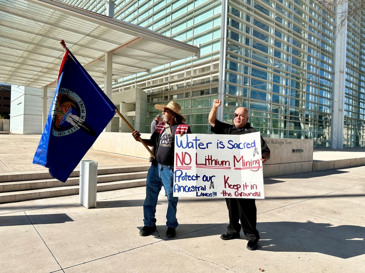 Two Hualapail Tribe members protest outside a courthouse, one holding a flag and another with a sign that says "Water is sacred, no lithium mining, protect our ancestral lands, keep it in the grounds"