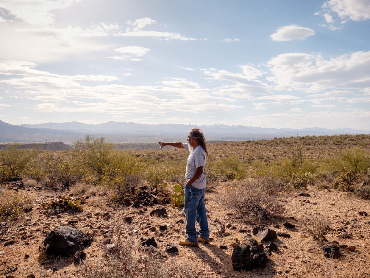 A man in a white T-shirt and jeans stands in a desert surrounded by rocks, pointing toward mountains in the distance.