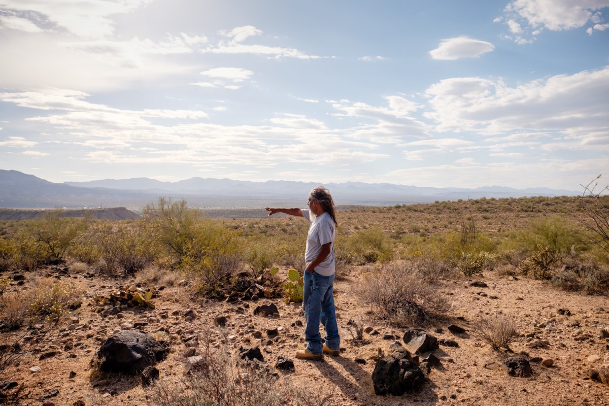 A man in a white T-shirt and jeans stands in a desert surrounded by rocks, pointing toward mountains in the distance.