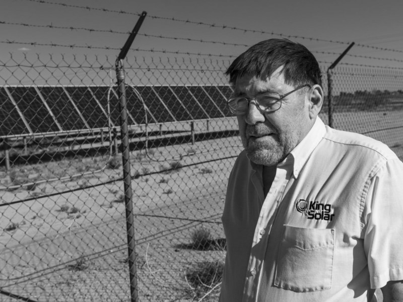 A man in a solar company shirt stands in front a barbed wire-surrounded solar array.