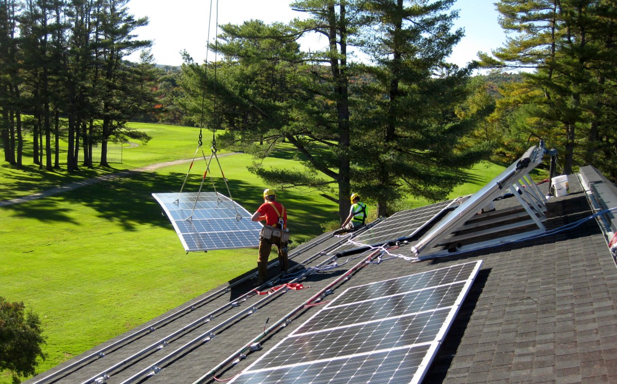 A crane lifts a solar panel onto a sloped roof as two workers await.