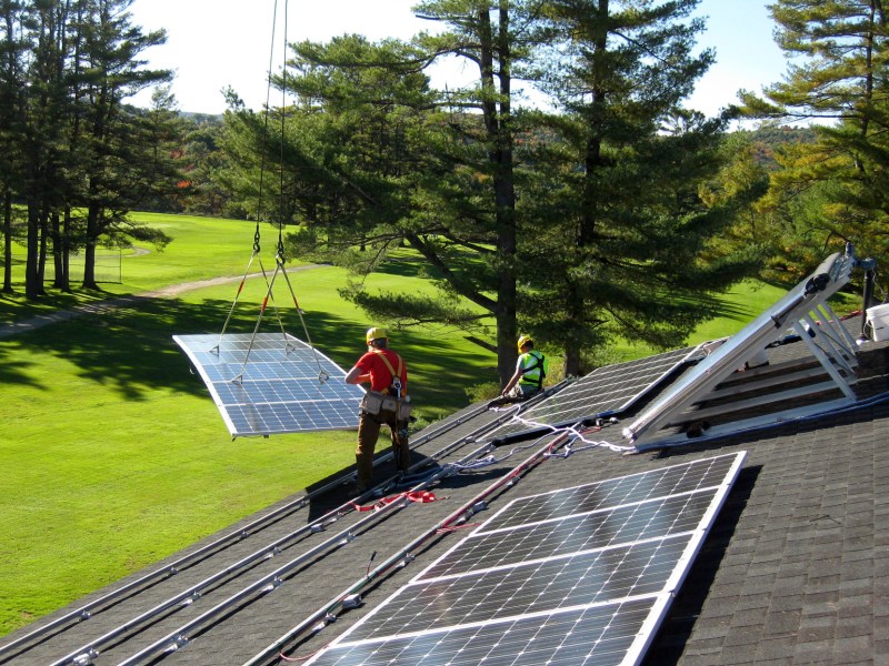 A crane lifts a solar panel onto a sloped roof as two workers await.