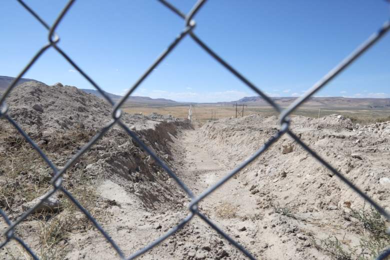 A deep trench in the desert is viewed through a chain-link fence.