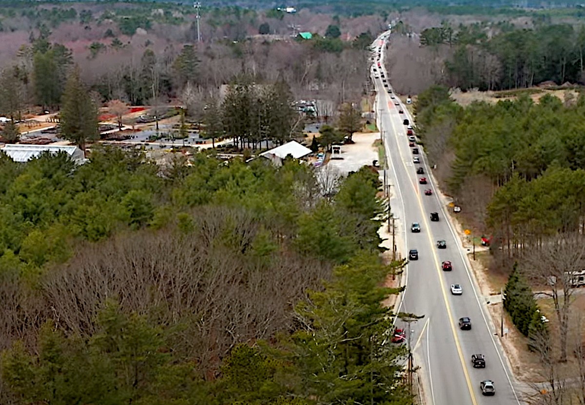 A video still showing heavy traffic on a two-lane highway through a wooded area of Maine that also features homes and commercial development.