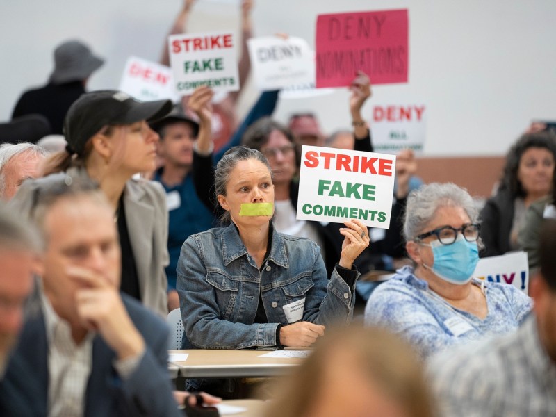 A group of protesters at a meeting. One protester with tape covering their mouth holds a sign that reads "Strike fake comments."