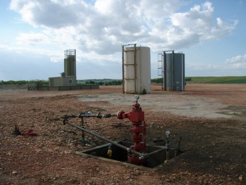 A fracking well head pokes out of a hole in the ground with large containers behind it